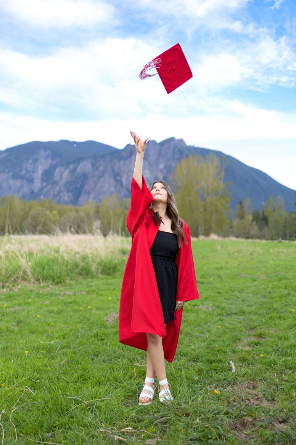 Young woman in red graduation gown tossing cap in air outdoors with mountain backdrop on a sunny day.