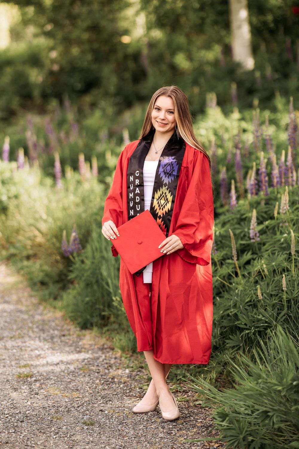 Young woman in red graduation gown and stole smiling outdoors among purple wildflowers on a garden path.