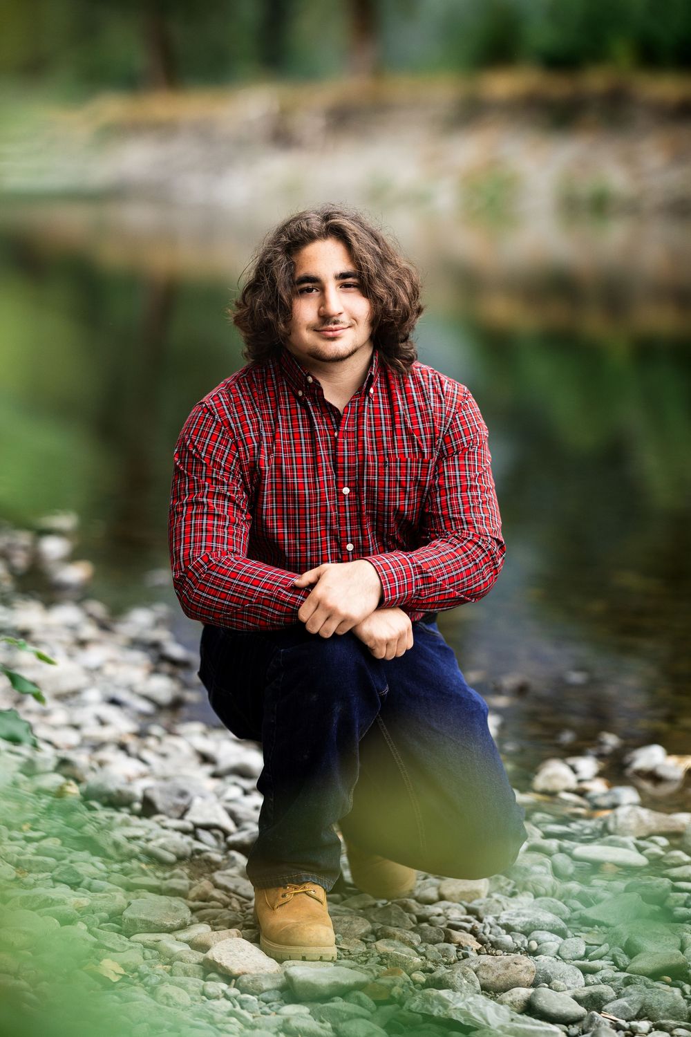Young man with curly hair in red plaid shirt crouching on rocky riverbank, smiling at camera.
