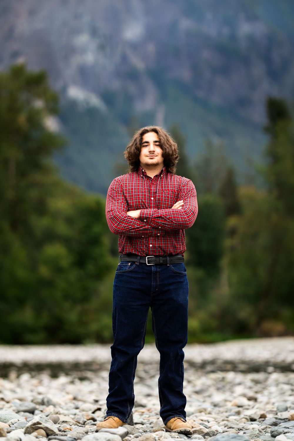 Young man with curly hair in red plaid shirt stands arms crossed on rocky terrain with mountain forest backdrop.