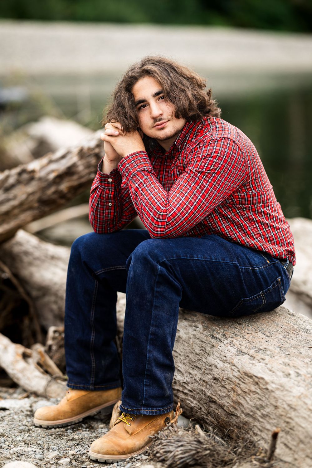 Young man with curly hair in red plaid shirt and jeans sits on driftwood logs near a lake.