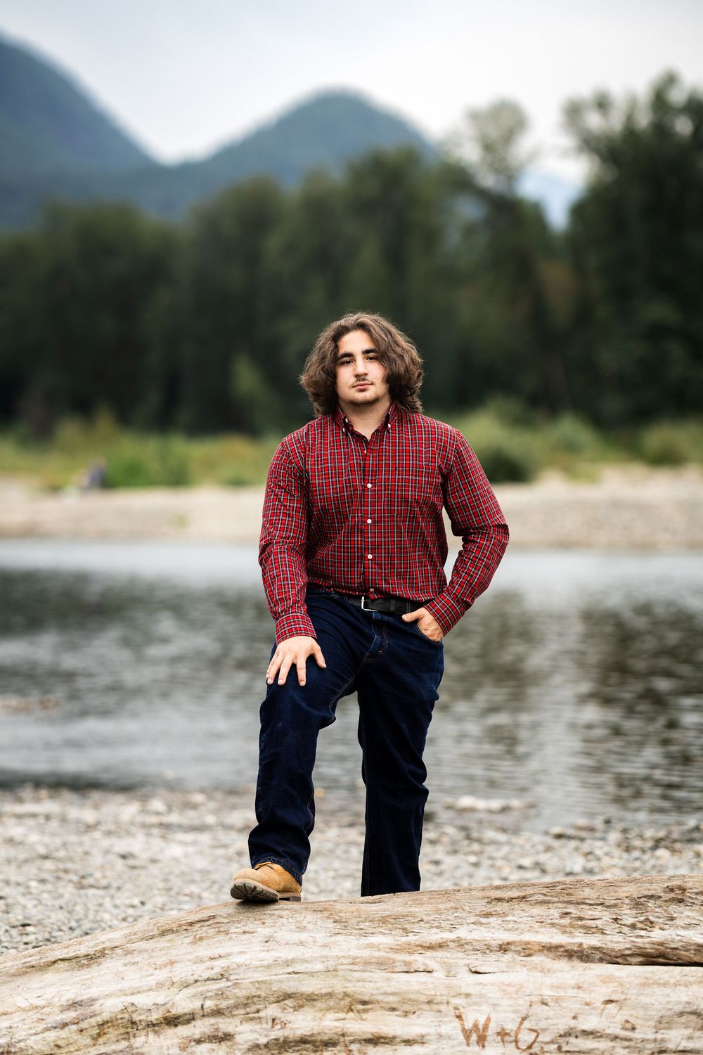 Young man with curly hair in red plaid shirt and jeans standing on rocks near a river with mountains behind.