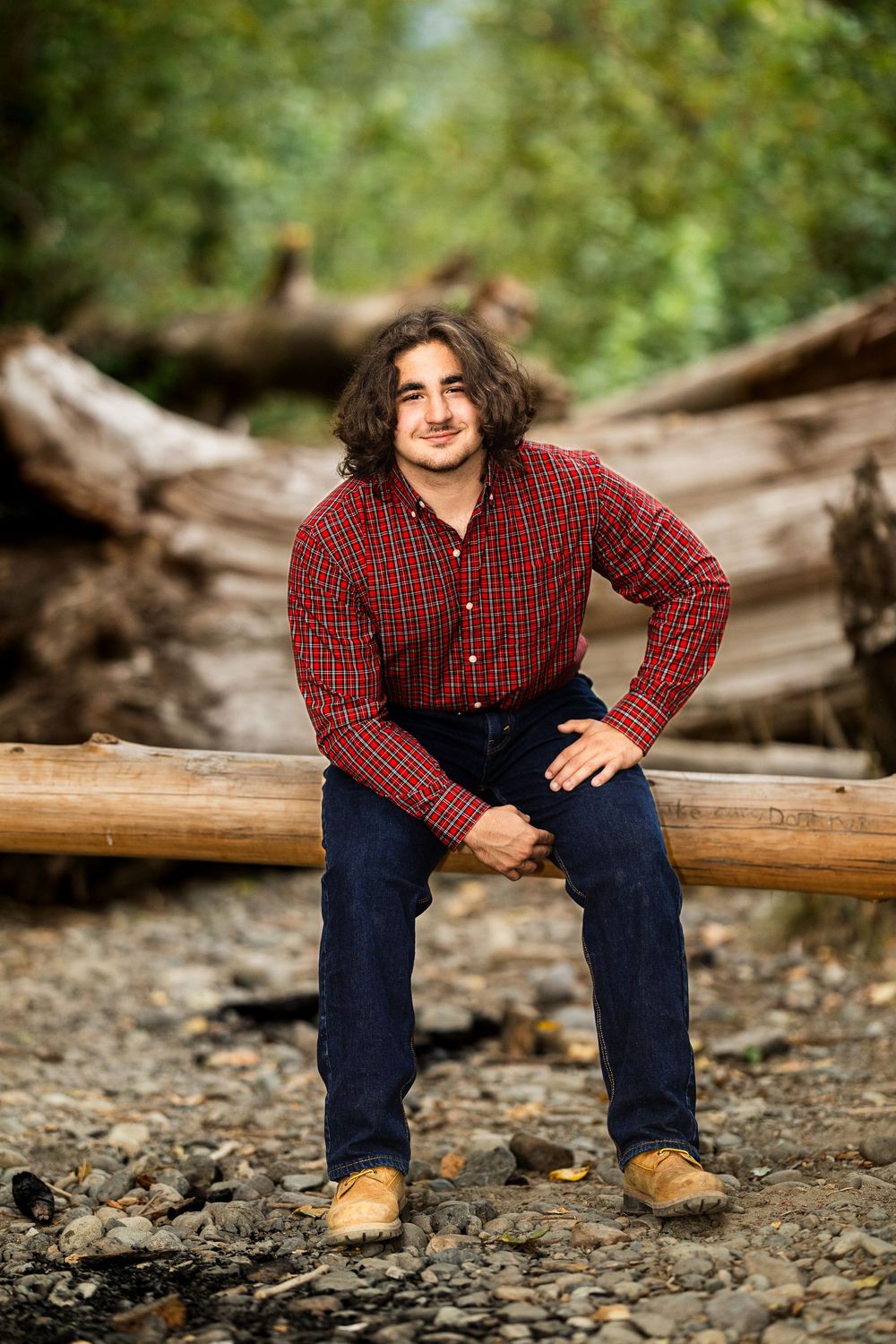 Young man with curly hair wearing red plaid shirt and jeans sitting on a log in a rocky forest setting.