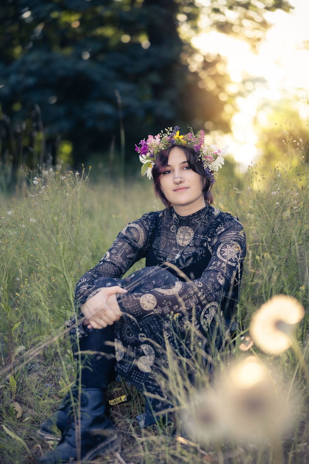 Young woman in black lace dress and floral crown sits in tall grass with golden backlight in nature.