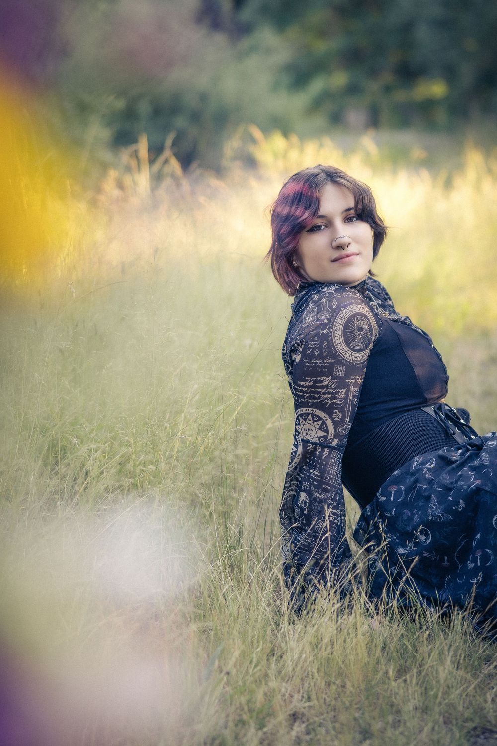 Woman in black lace dress sitting in tall grass field, smiling over shoulder in soft natural light.