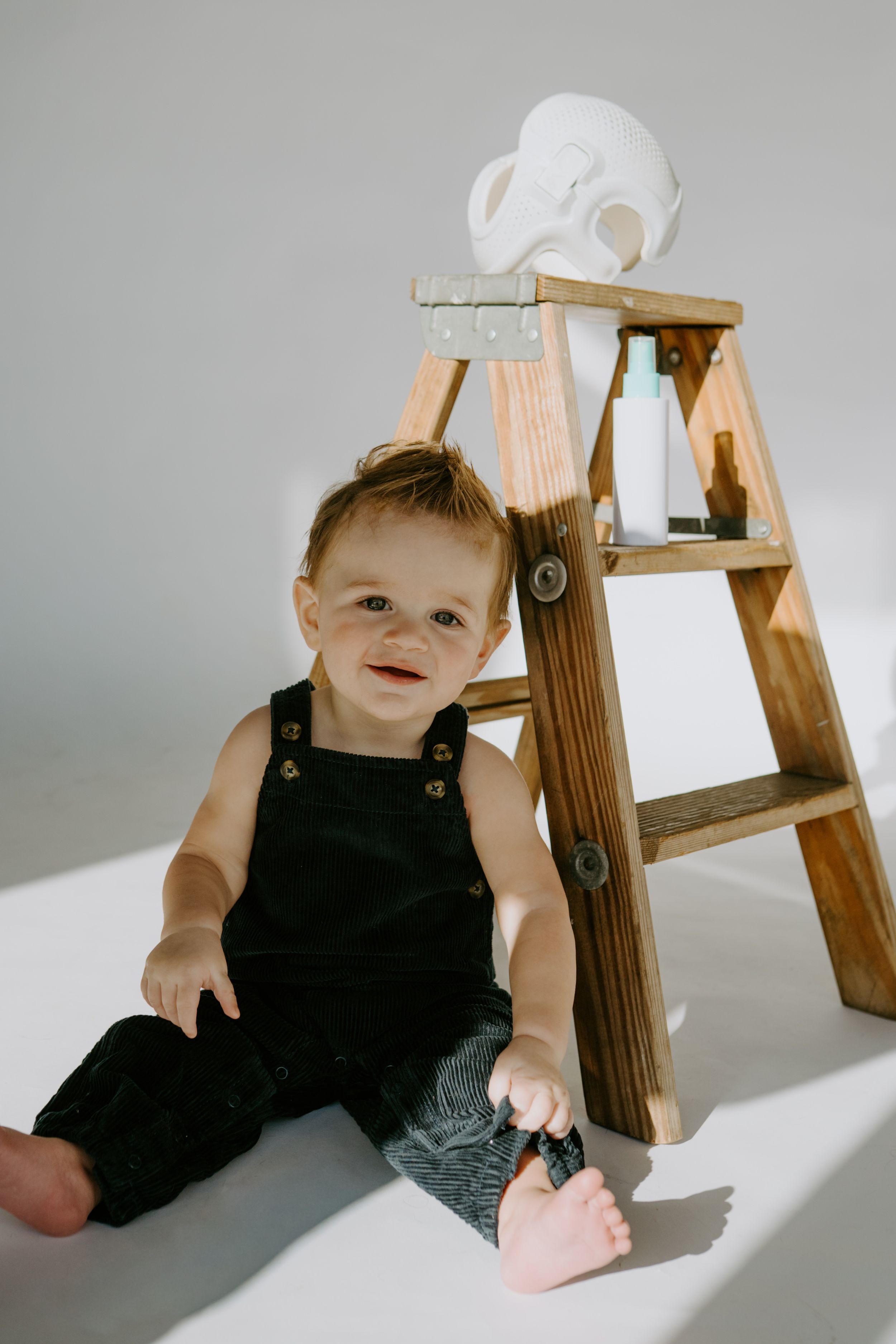 Small child in black overalls sitting beside wooden ladder with white mask on top.