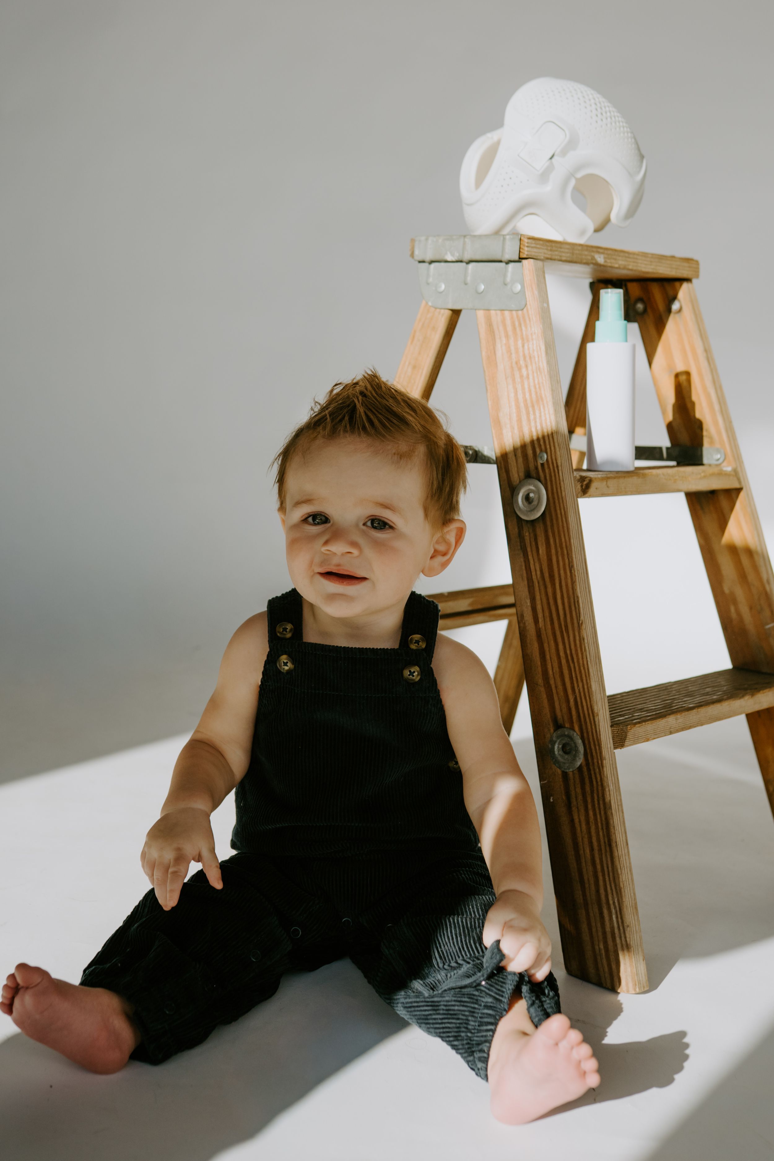 Small child in black overalls sitting beside wooden ladder with white helmet.