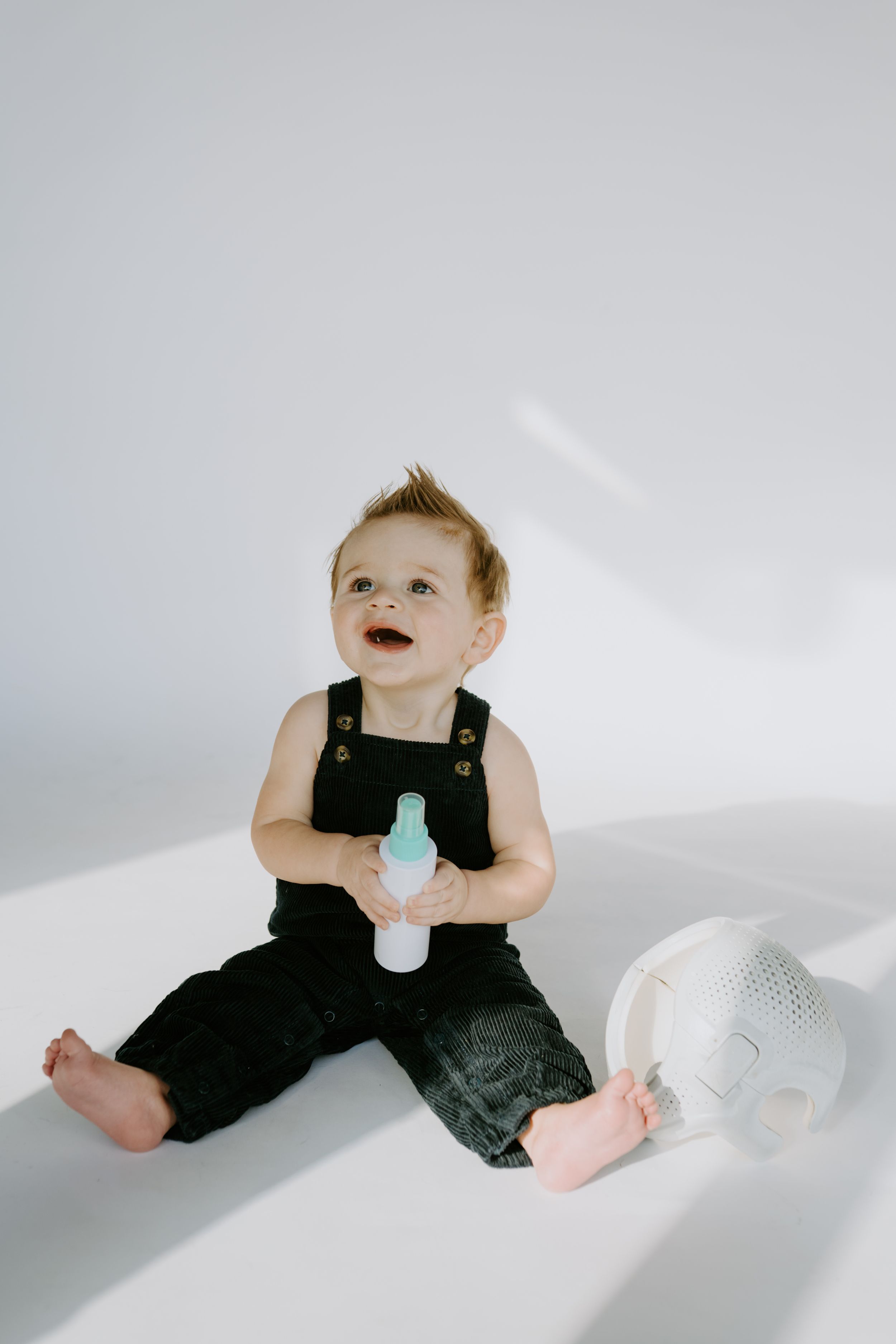 Small child in black overalls holding spray bottle on white background.