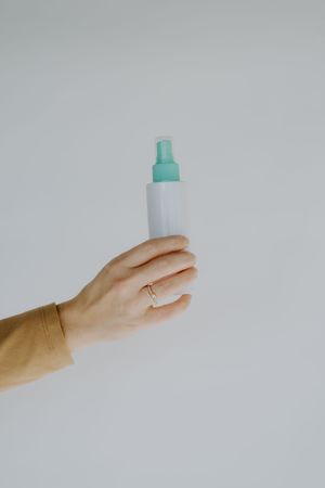 Hand wearing ring holds white spray bottle with turquoise cap against white background.