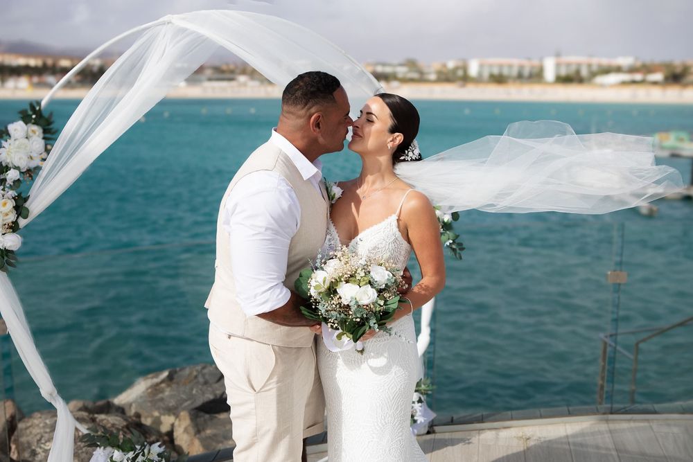 Bride and groom share a kiss by turquoise ocean, veil flowing in breeze, bride holding white floral bouquet.