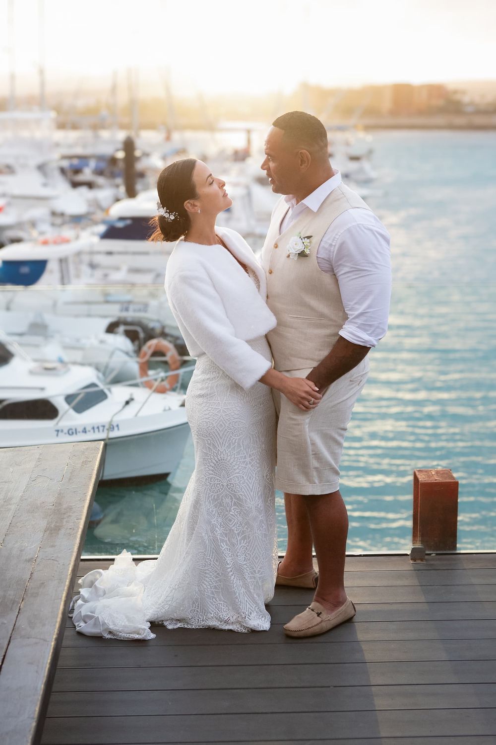 Bride and groom in white attire share a romantic moment on a marina dock at sunset with boats in the background.