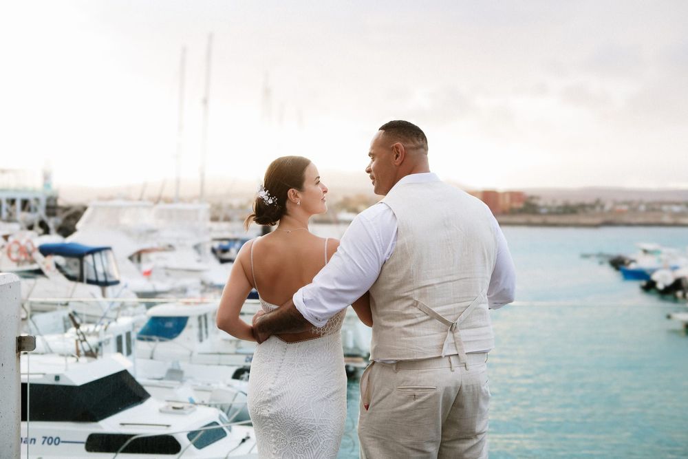 Bride and groom stand together overlooking a marina with boats and calm water at sunset.