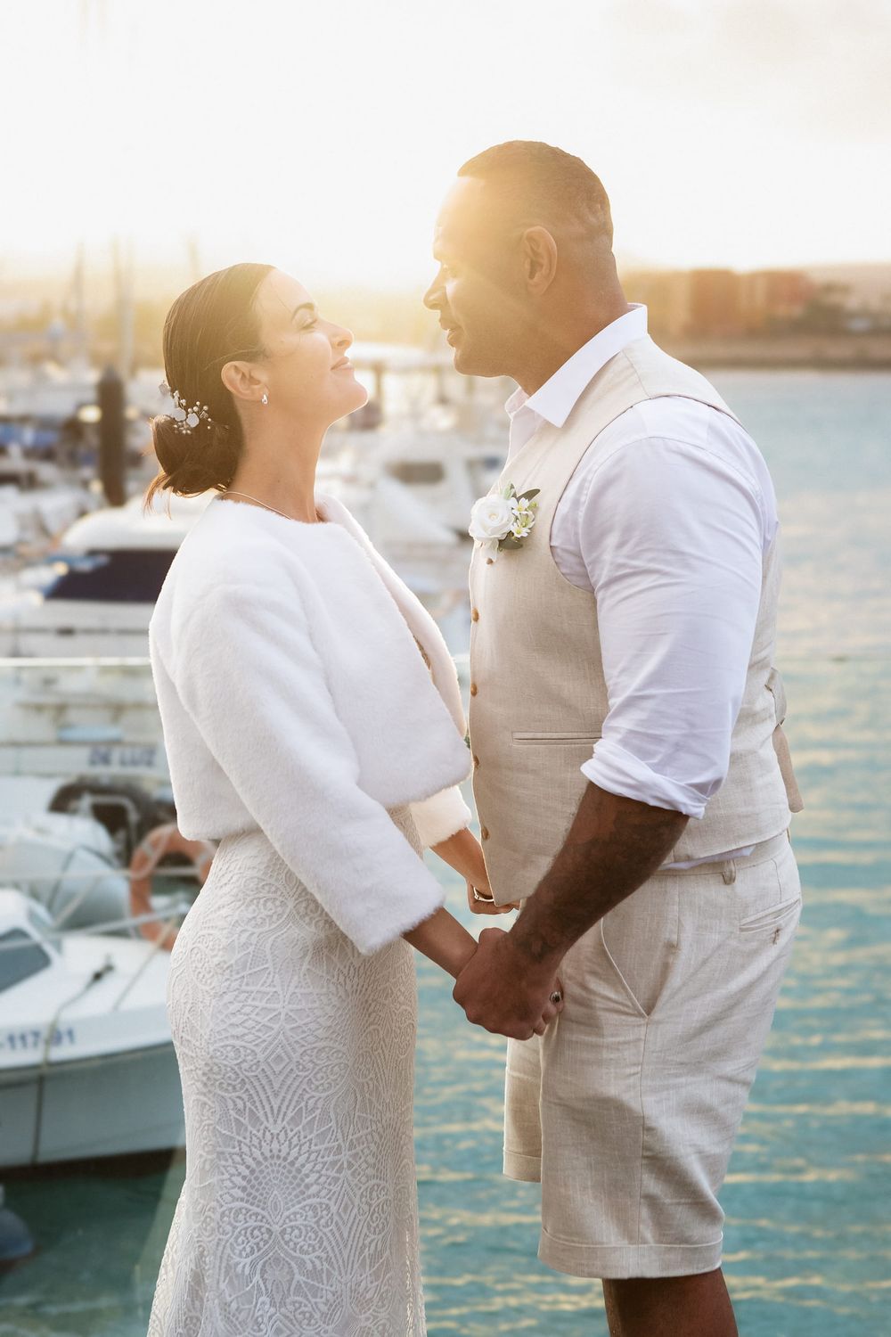 Couple in white wedding attire holding hands and smiling at sunset by a marina with boats in background.