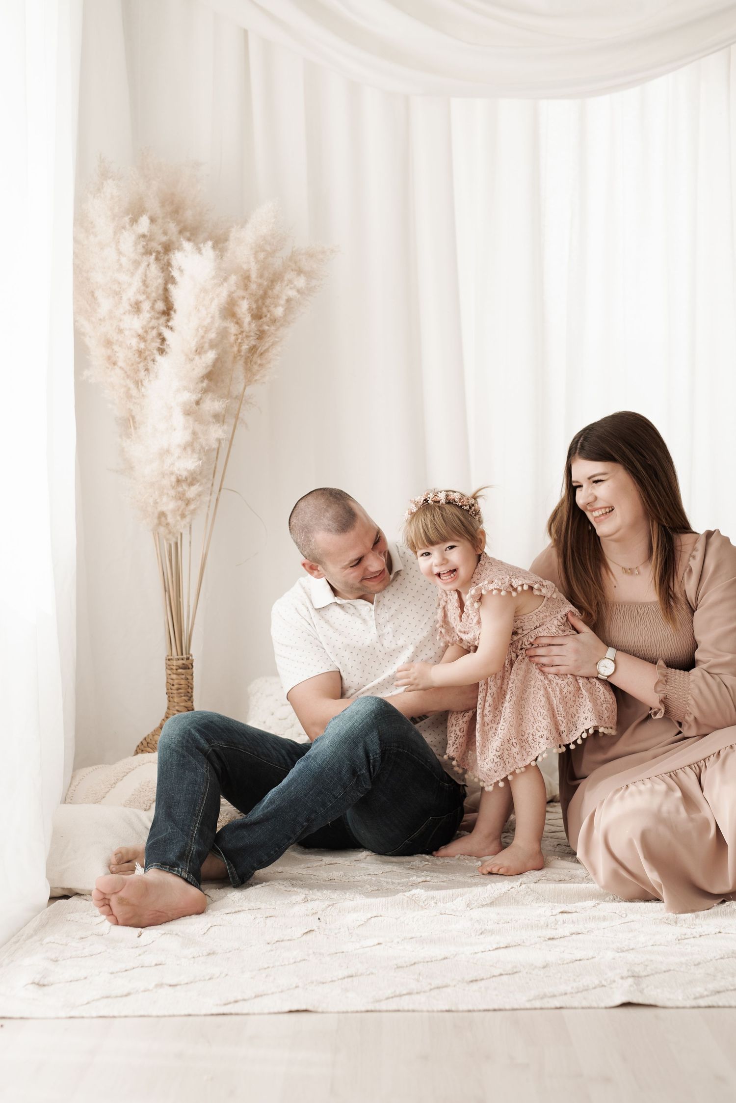 Happy family moment captured in soft neutral tones against white backdrop with pampas grass decor.