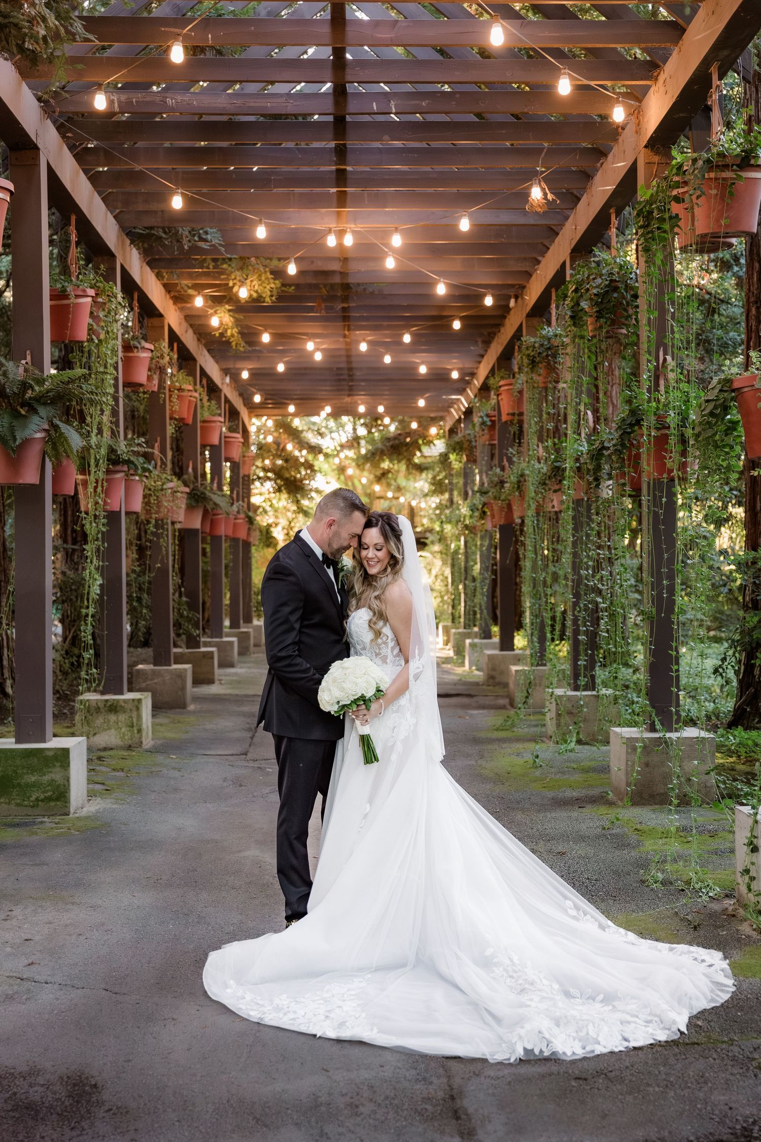 A romantic wedding photo in a covered walkway with string lights and hanging plants creating a magical garden atmosphere.