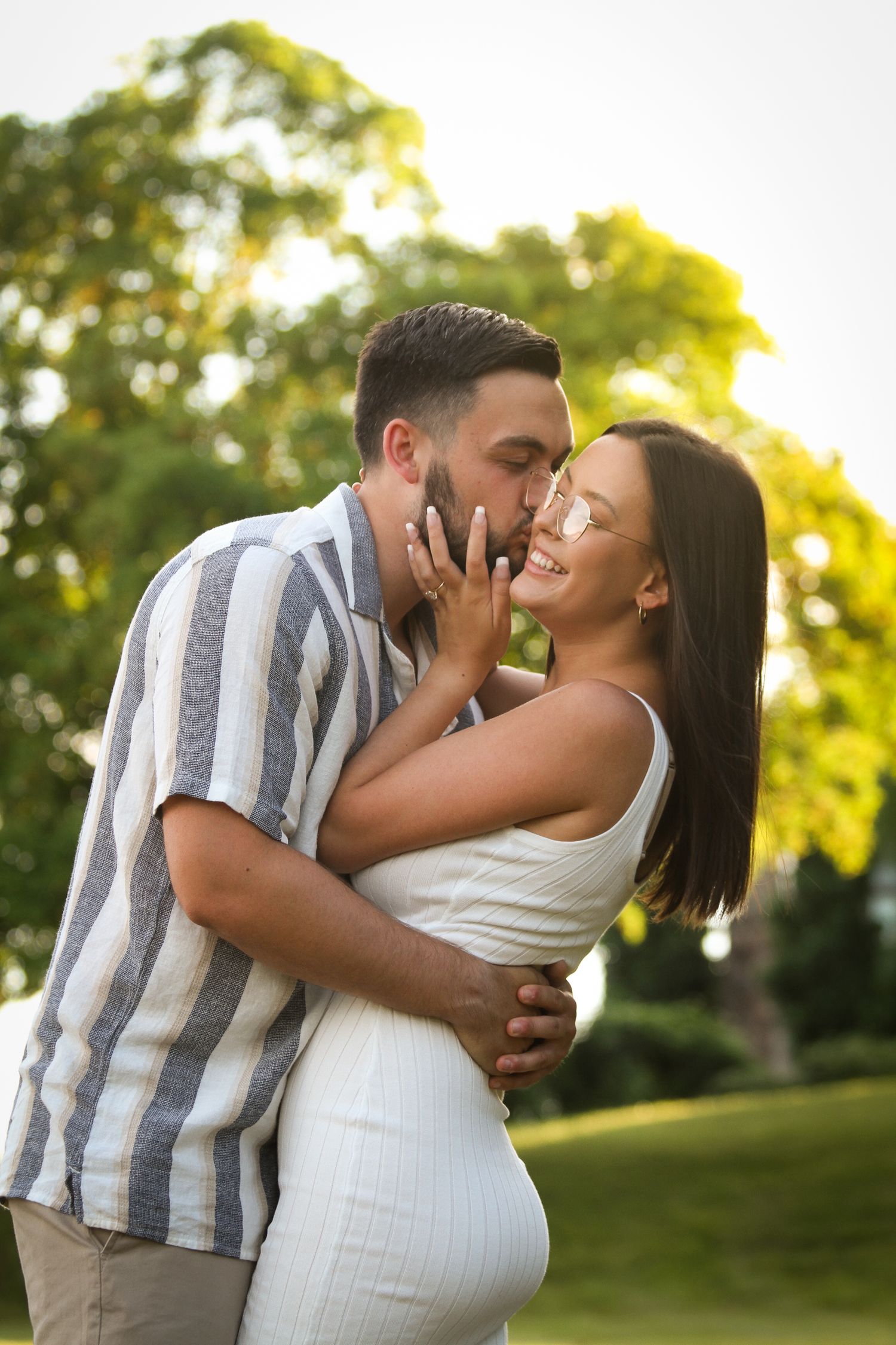 A man leans in to kiss his fiance during their summer engagement photo session in a Kansas City park at golden hour.