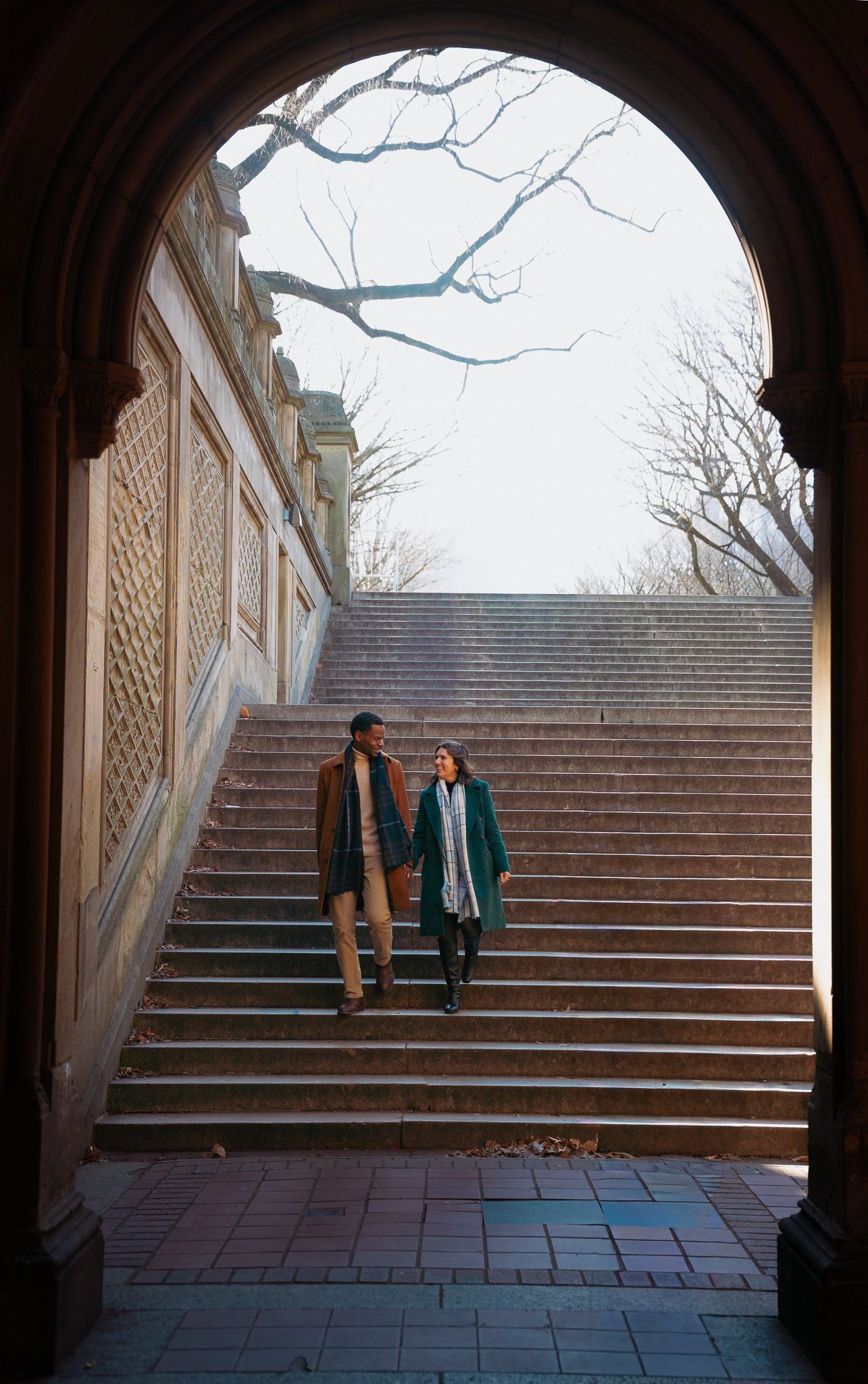 A couple descending stone steps framed by an architectural archway on a sunny day.