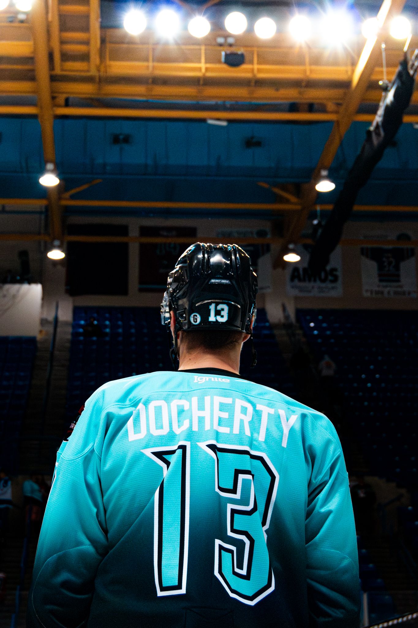 Hockey player wearing teal jersey number 13 with 'Docherty' nameplate stands under arena lights.