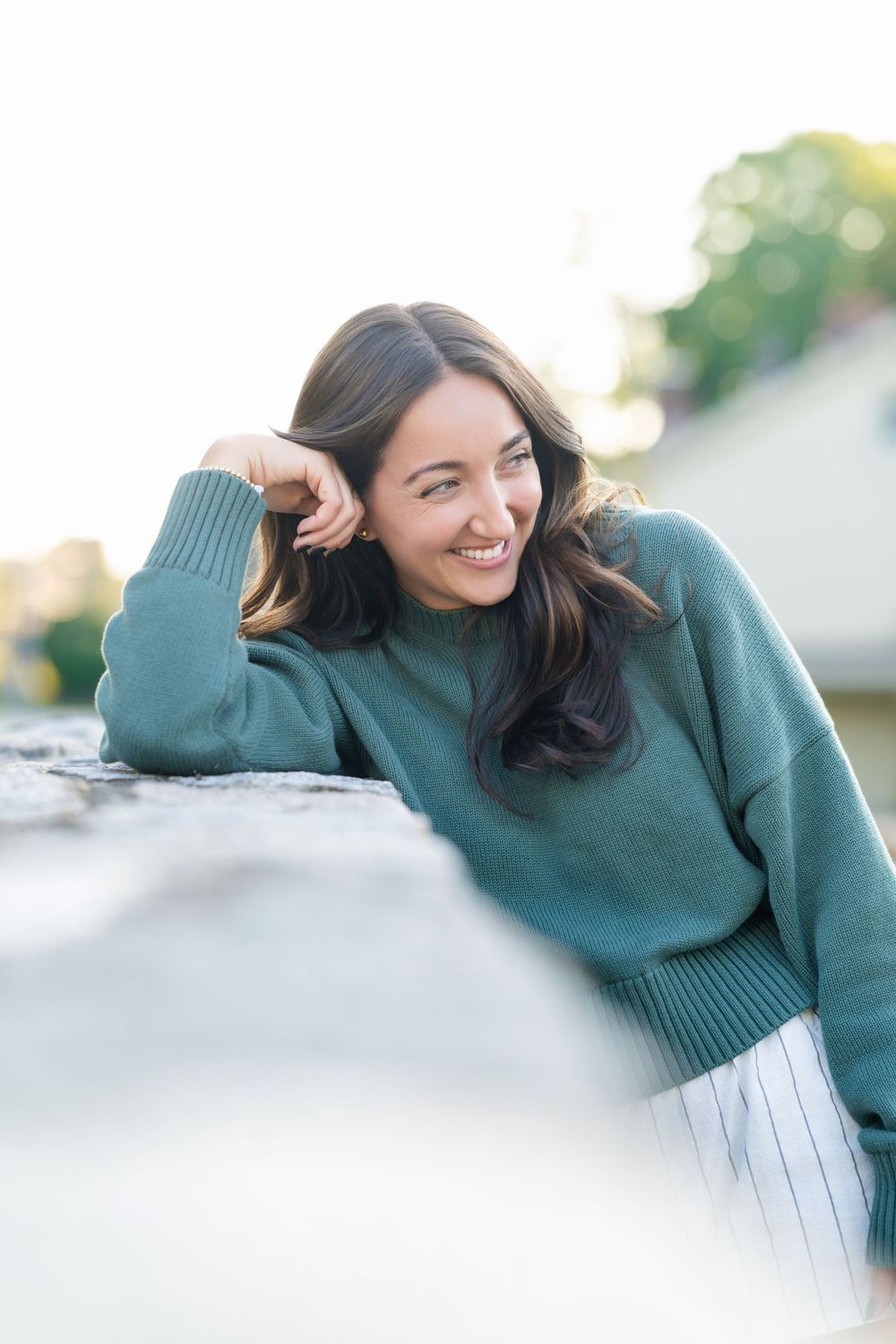 Woman in green sweater smiling and touching hair in soft natural lighting.