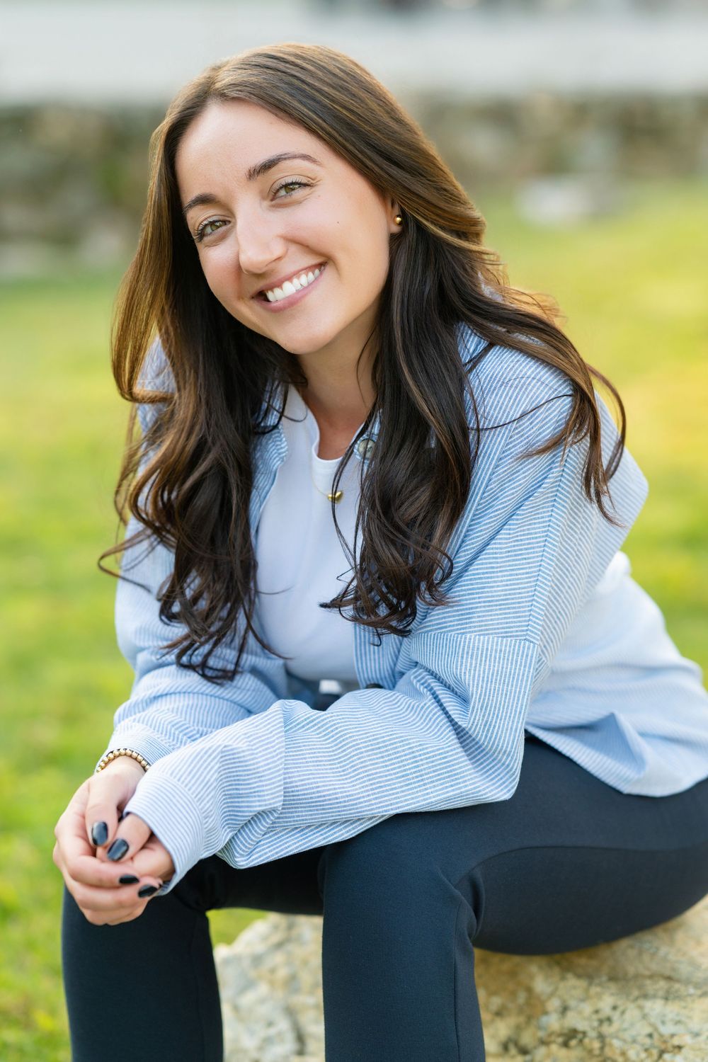 Young woman in a light blue sweater sits outdoors on a rock with a warm smile.
