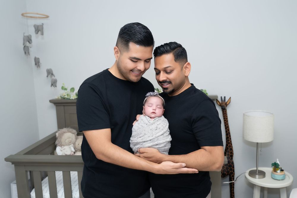 Two people in black shirts share tender moments with a baby while sitting on a grey couch in a modern living room.