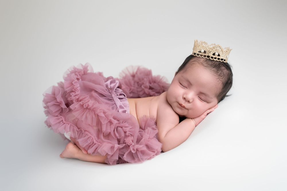 A sleeping newborn wearing a pink tutu and tiara rests peacefully on a white backdrop in a delicate pose.