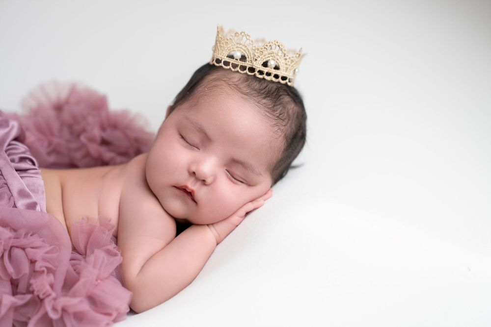 Sleeping newborn baby wearing a delicate crown lies peacefully on pink tulle fabric in a professional portrait setting.