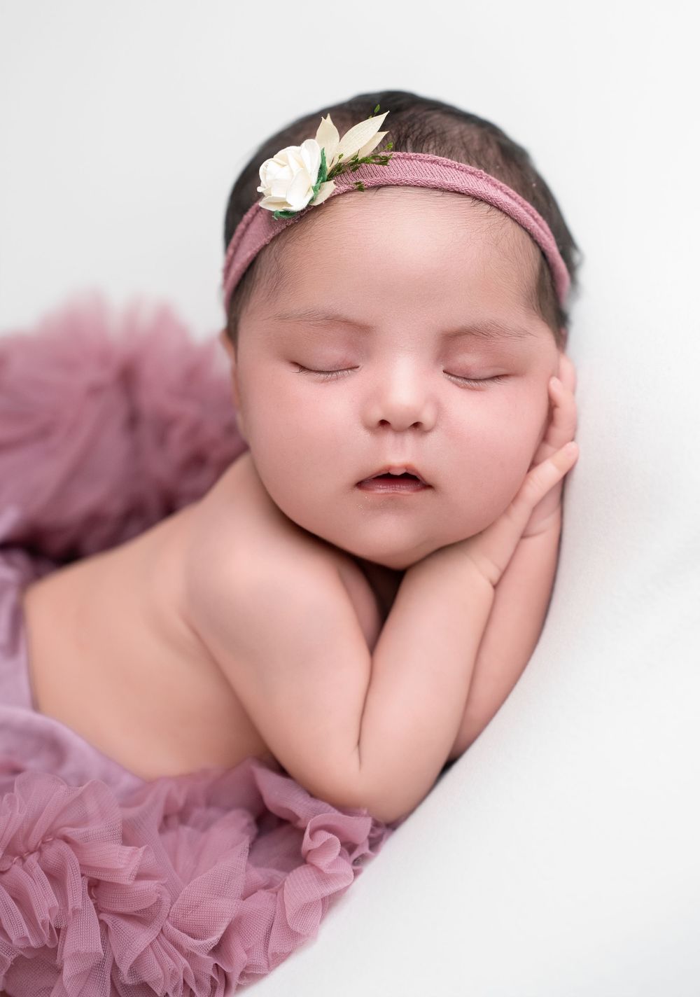 Sleeping newborn wearing pink tulle dress and delicate floral headband rests peacefully on white backdrop.