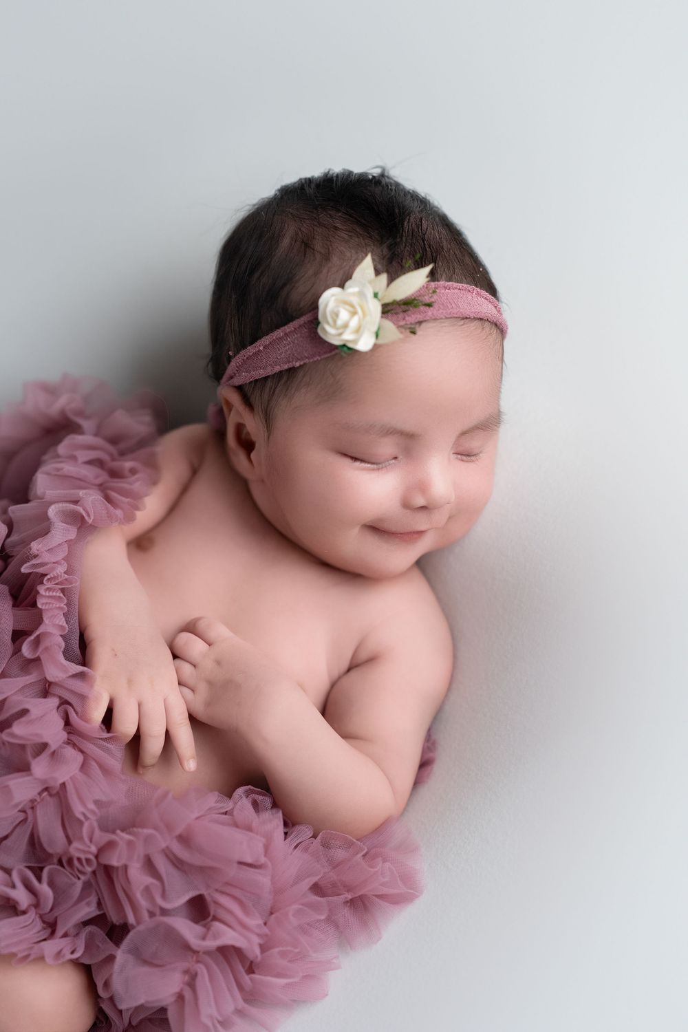 Sleeping newborn baby wearing a delicate white hair bow and pink tulle wrap in soft studio lighting.