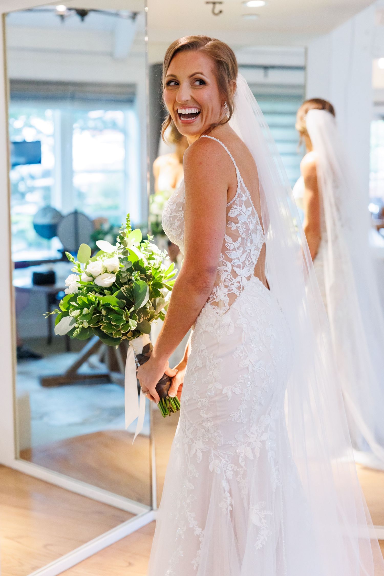 A bride in a beautiful lace wedding dress laughs joyfully while holding a white and green bouquet in a bright room at Big Cedar Lodge.