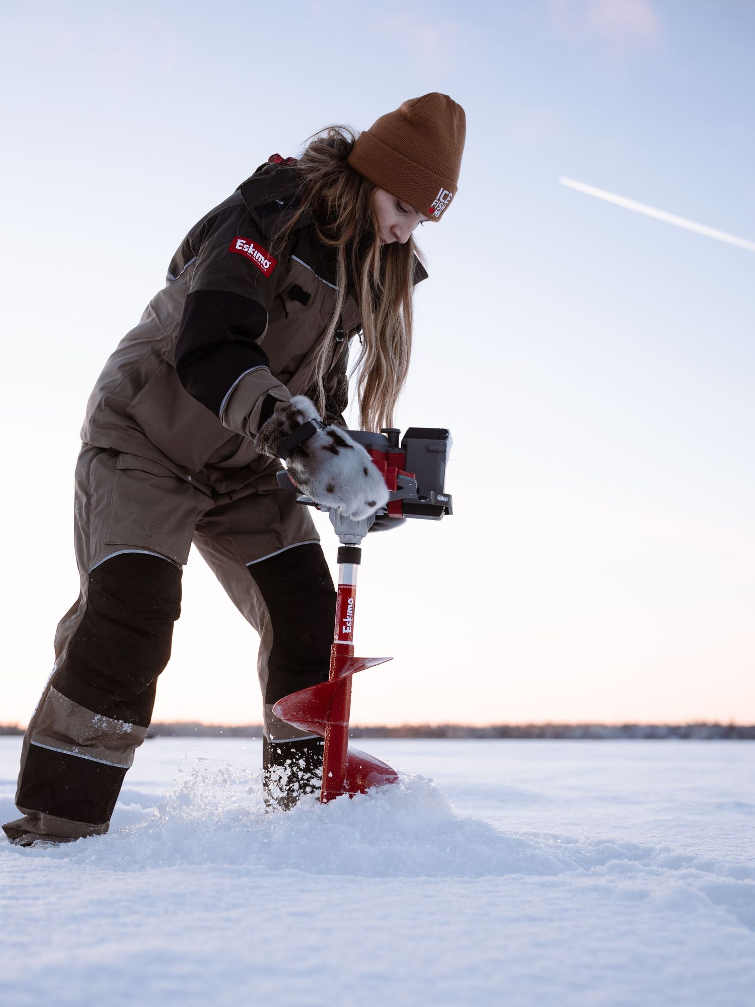 Winter ice fishing scene with person drilling hole using red power auger on frozen lake.