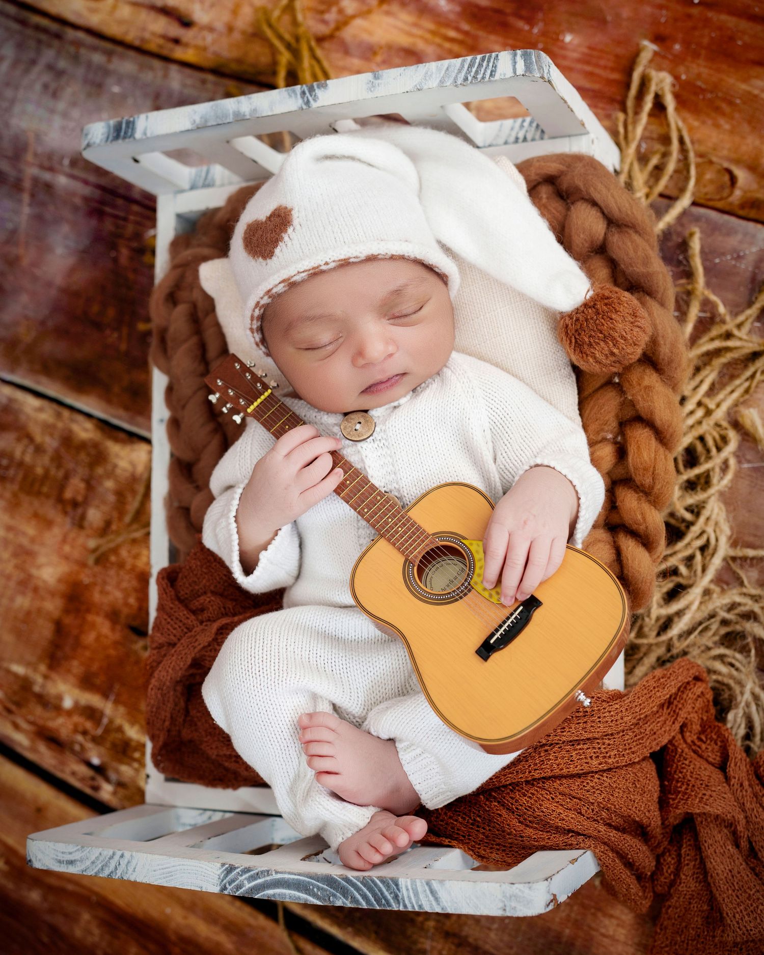 Sleeping baby in white outfit lying on brown blanket with miniature acoustic guitar prop in rustic setting.