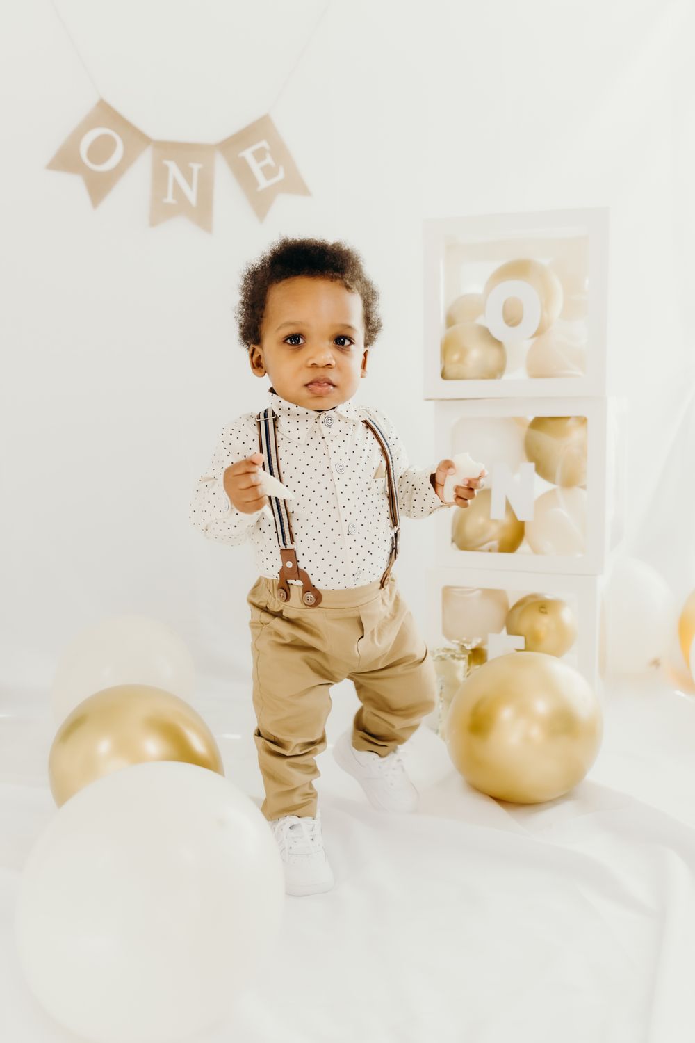Baby in polka dot shirt and khaki pants poses with gold balloons and decorations for first birthday.