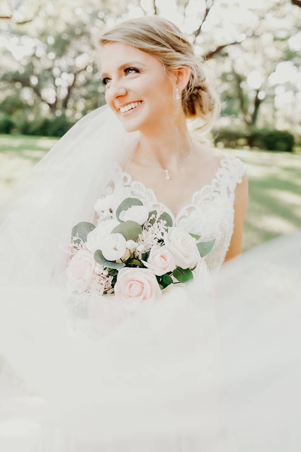 A radiant bride holds a soft pink and white bouquet while wearing a flowing veil outdoors.
