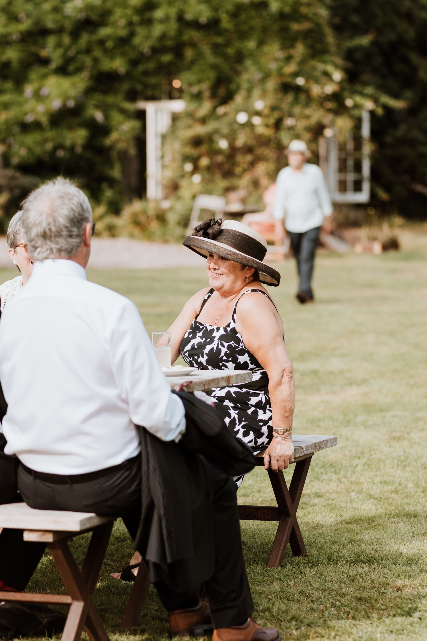 Guests sit on wooden benches at an outdoor wedding reception on a sunny day.