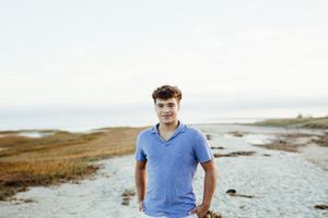 Young man in blue shirt standing on coastal shoreline with grassy marshland and water in background.