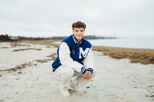 A young boy in a navy sweater sits on a sandy beach near the ocean on an overcast day.