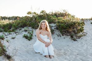 A person in a white dress sits on a sandy beach near green coastal vegetation during golden hour.