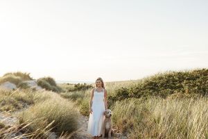 A woman in a white dress walks through tall grass in a golden field at sunset.