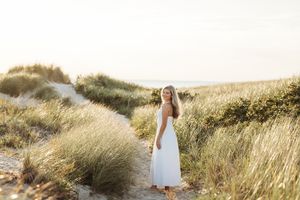 A person in a flowing white dress walks through tall golden grass in a coastal meadow at sunset.