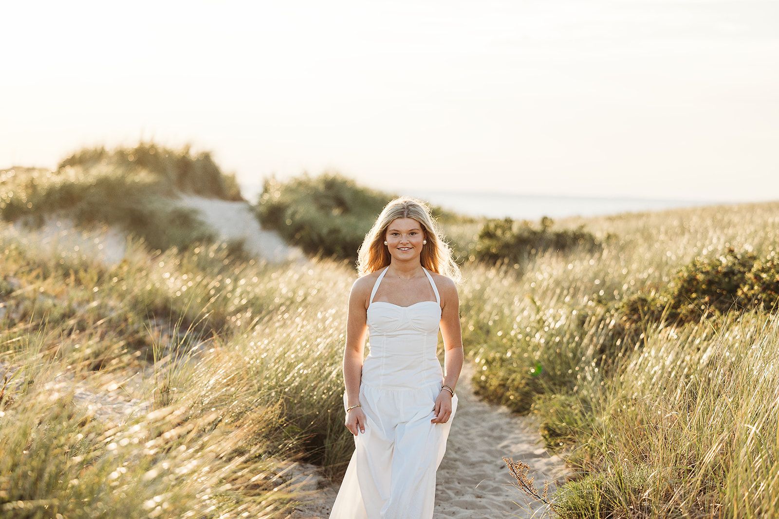 A person in a flowing white dress walks through a golden field of tall grass at sunset.