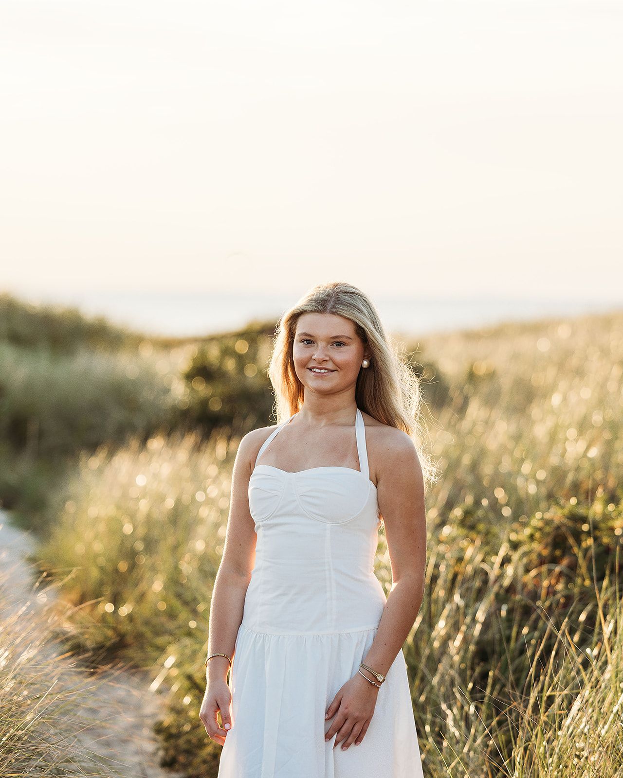 A woman in a white dress stands in a sunlit field of tall grass with a soft, warm glow in the background.