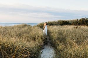 A sandy path through tall grass leads to a distant beach and ocean horizon on a cloudy day.