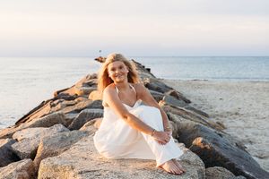 Woman in white dress sitting on rocky coastal jetty at sunset with ocean in background.