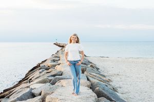 Woman in white shirt and jeans walks on rocky jetty extending into calm ocean under overcast sky.