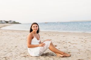 A person in a white dress sits on a sandy beach with ocean waves and coastline in the background.