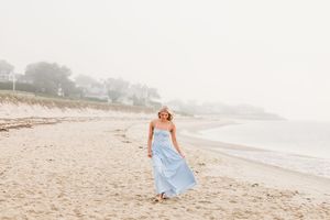 A person in a light blue dress walks along a misty beach shoreline at sunrise.