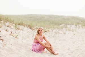 A person in a pink dress sits on a sandy beach dune while looking towards the ocean at sunset.