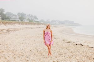 A person in a pink dress walks barefoot along a misty beach shoreline on an overcast summer day.