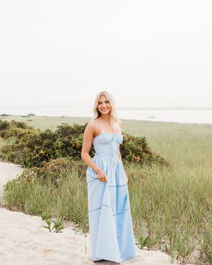 Woman in light blue summer dress poses on sandy beach path with coastal vegetation in background.